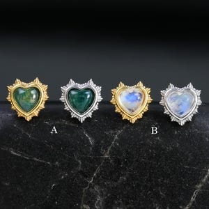 Op de afbeelding: Vier hartvormige oorstekers met edelsteen centra. De oorbellen zijn gezet in goud en zilver metaal met een decoratieve rand. De edelstenen omvatten groen en blauw getinte stenen. De oorbellen worden getoond op een zwarte steen.