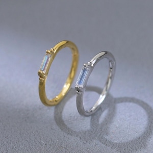 May include: Two slender rings, one gold and one silver, each adorned with a rectangular, iridescent gemstone. The rings are displayed against a muted background, emphasising their understated charm. The gold ring has a warm, polished finish, while the silver ring has a cool, reflective sheen.