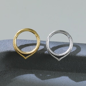 Può includere: Due anelli per septum, uno dorato e uno argentato, sono esposti su una superficie grigio scuro. Ogni anello presenta un bordo decorativo con perline e una punta inferiore. Gli anelli sono progettati per piercing al naso.