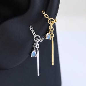 Può includere: Due orecchini a catena pendenti, uno color argento e uno color oro, ciascuno caratterizzato da una piccola pietra azzurra a forma di goccia e da un sottile ciondolo a barra metallica dritta. Presentati su un supporto nero a forma di orecchio.