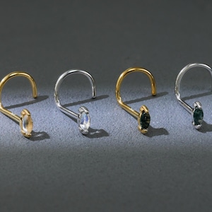 May include: Four nose rings with curved posts. Two are gold-toned, one is silver-toned, and one is a different silver tone. Each has a small, oval-shaped gemstone in various colors: yellow, white, and two shades of green. The jewelry is displayed on a gray surface.