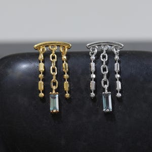 Puede incluir: Dos pendientes colgantes, uno dorado y otro plateado, cada uno con una barra superior curva y tres cadenas colgantes. La cadena central de cada pendiente sostiene una pequeña piedra preciosa rectangular de color azul claro.