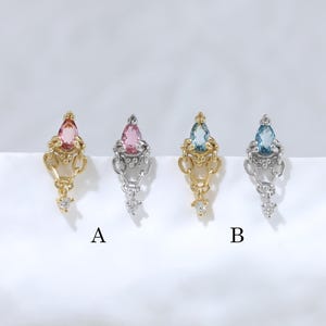 Peut inclure: Quatre breloques décoratives en métal disposées en rangée, avec des pierres en forme de goutte d'eau roses et bleues. Deux breloques sont dorées et deux sont argentées, ornées de petites pierres claires. Les lettres A et B sont imprimées.
