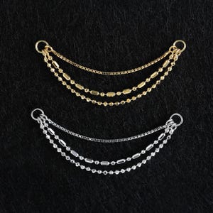 Op de afbeelding: Twee sieraden, één goudkleurig en één zilverkleurig, elk met drie delicate kettingen. De bovenste ketting is een dunne, gladde streng, terwijl de onderste twee kleine, getextureerde kralen hebben. Elk stuk heeft een kleine lus aan de bovenkant.