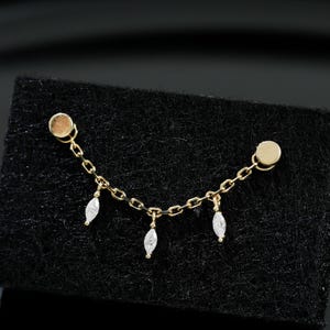 May include: Gold-coloured earring featuring a chain with three hanging, teardrop-shaped white gemstones. The earring has two round posts, one at each end of the chain. The earring is displayed on a black surface.