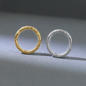 May include: Two small, round earrings. One is gold-colored, the other is silver-colored. Both earrings have a textured, patterned design around the outer edge. The earrings are displayed on a gray surface.