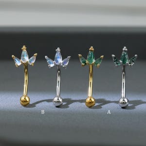 Może przedstawiać: Cztery zakrzywione kolczyki do brwi. Dwa są w kolorze złotym z małą złotą kulką na dole i koroną z niebiesko-zielonych kamieni szlachetnych. Pozostałe dwa są w kolorze srebrnym z małą srebrną kulką na dole i koroną z niebiesko-zielonych kamieni szlachetnych.