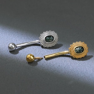 Peut inclure: Deux piercings de nombril. L'un est argenté, l'autre doré. Chacun possède une petite pierre ovale vert foncé entourée d'un motif texturé et perlé. Le piercing argenté est complet, tandis que le piercing doré est partiellement démonté.