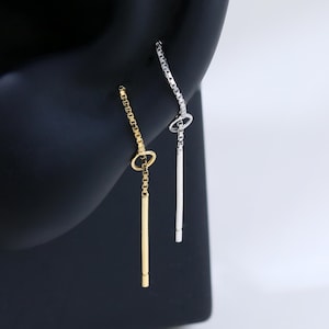 May include: Two minimalist earrings, one gold and one silver, each with a small circle and a dangling bar. The earrings are set against a black backdrop, highlighting their simple design and elegant appearance.