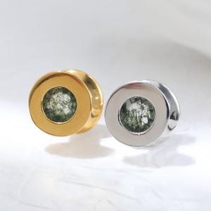 Op de afbeelding: Twee ronde oorplugs, één van goudkleurig metaal en één van zilverkleurig metaal. Elke plug heeft een centrale inleg van mosagaat, een doorschijnende steen met donkergroene, takachtige insluitingen zichtbaar binnen het ronde frame.