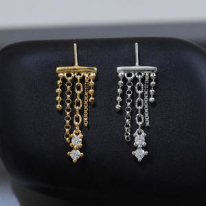 May include: Two dangle earrings, one gold-toned and one silver-toned, each featuring a horizontal bar post with multiple hanging chains of varying textures and two small, clear, round-cut stones at the bottom of the central chain.