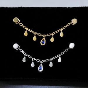 Peut inclure: Deux paires de boucles d'oreilles pendantes. Une paire est dorée avec des breloques en forme de goutte et une gemme bleue. L'autre paire est argentée avec des breloques en forme de goutte et une gemme bleue. Les deux paires sont présentées sur une surface noire.