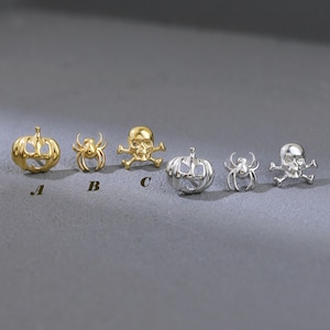 May include: A collection of Halloween-themed earrings in gold and silver tones. Designs include pumpkins, spiders, and skulls with crossbones. The earrings are displayed on a gray surface, showcasing their intricate details and festive appeal.