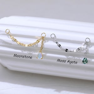 May include: Two ear chains, one gold-toned and one silver-toned, each with a chain of small, flat links. The gold chain has a pear-shaped moonstone, and the silver chain has a green moss agate. Text on the image reads "Moonstone" and "Moss Agate."
