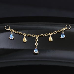 Pode incluir: Uma peça de conexão de corrente dourada com cinco pingentes de pedra da lua em forma de gota. O pingente central pende de um segmento curto de corrente vertical, enquanto os outros quatro estão espaçados uniformemente ao longo da corrente.