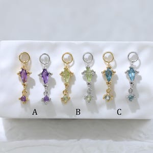 May include: Six vertical jewelry charms arranged in a row on a white textured block. Each charm features a kite-shaped gemstone topped with a circular jump ring and a small dangling round stone. The charms alternate between gold and silver metal tones.