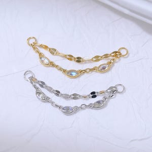 May include: Two pairs of delicate earrings. One pair is gold-toned, the other is silver-toned. Each earring features a chain of small, flat links and oval-shaped iridescent stones. The earrings are designed to be worn through multiple piercings.