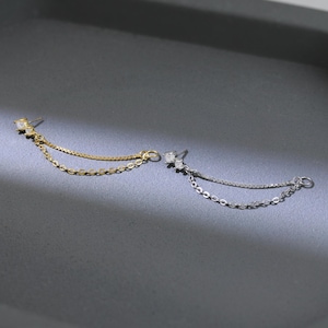 May include: Two delicate earrings with a chain design. One earring is gold-toned, the other is silver-toned. Each earring features a small, square-cut gemstone at the stud and a chain that dangles down. The earrings are displayed on a dark gray surface.