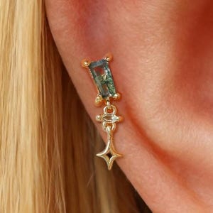 May include: A gold-colored earring with a rectangular green gemstone and a dangling star-shaped charm. The earring is attached to an ear, with blonde hair visible in the background. The gemstone has a unique, textured appearance.