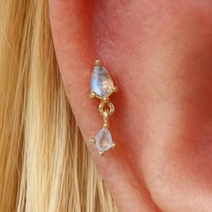 May include: A gold-colored earring with two teardrop-shaped gemstones. The top gemstone is a light blue color, and the bottom gemstone is clear. The earring is attached to the ear.