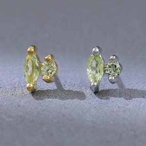 May include: A pair of stud earrings featuring a gold and silver finish. Each earring has a marquise-cut, light green gemstone paired with a smaller, round gemstone. The earrings are set against a gray background.