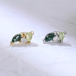 May include: Two delicate stud earrings. Each earring features a leaf design with green and white gemstones. One earring has a gold setting, the other a silver setting. The earrings are set against a white background.