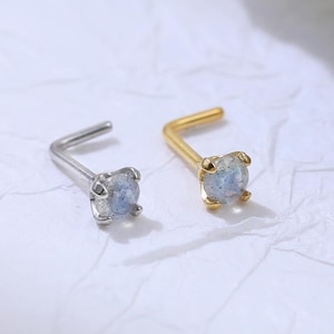 May include: Two nose studs, one silver-toned and one gold-toned, each featuring a square, light blue gemstone. The studs are L-shaped with a small, square gemstone at the end of the post. The background is a textured white surface.