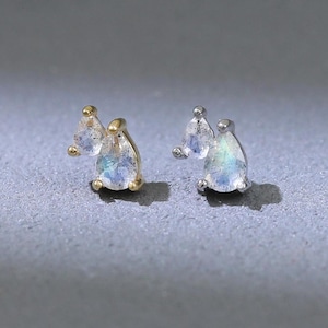 Puede incluir: Un par de pendientes de botón con piedras preciosas iridiscentes en forma de pera. Un pendiente es dorado, el otro plateado. Cada pendiente tiene un acento de piedra preciosa redondo más pequeño. Los pendientes se muestran sobre una superficie gris.