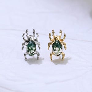 May include: Two insect-shaped earrings. One is silver-toned, the other gold-toned. Both have a green, oval-shaped stone in the centre. The earrings are on a white, textured surface.