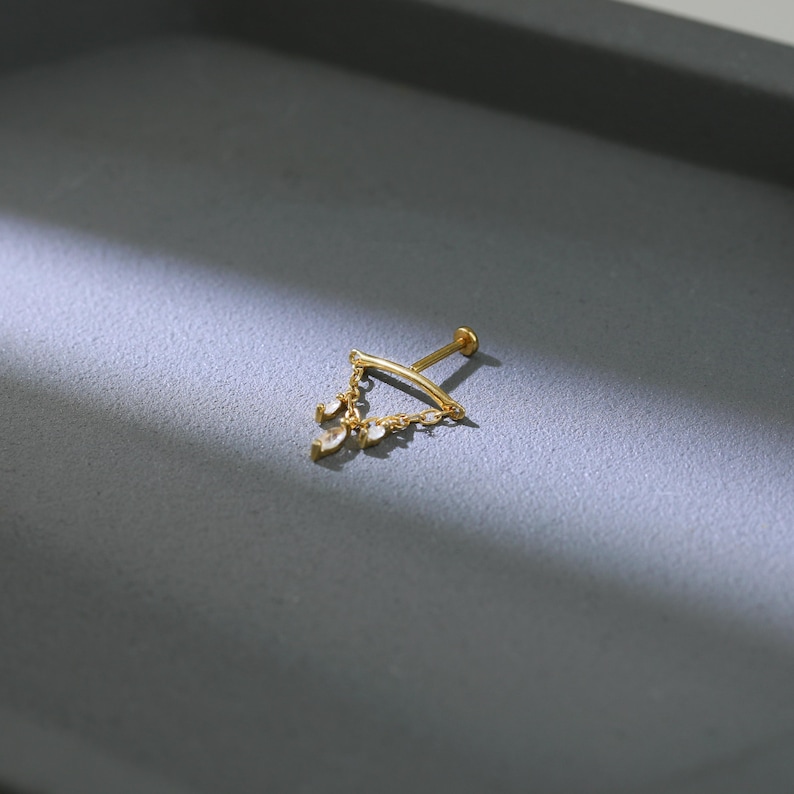 May include: A gold-colored lip piercing with a triangular design. The piercing features a curved bar with a flat disc on one end and a threaded post on the other. The triangle is adorned with small, dangling, teardrop-shaped gems.