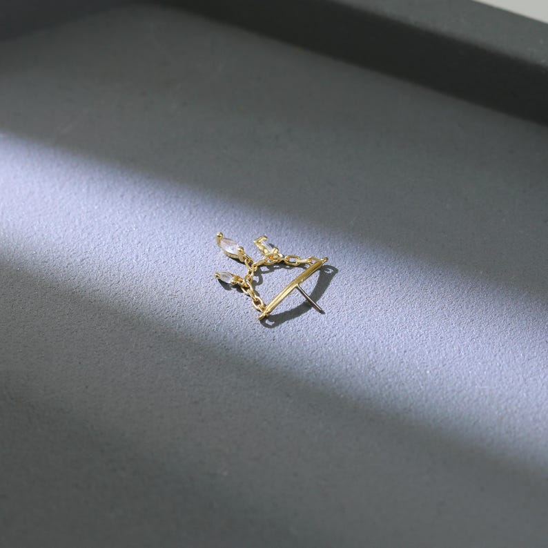 May include: A gold-colored earring with a delicate design. The earring features a series of small, clear, teardrop-shaped stones connected by fine chains. The earring post is visible, and the overall design is elegant and minimalist.