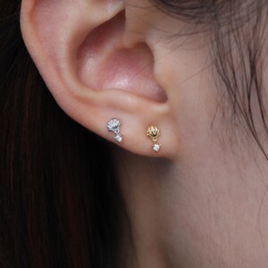 Puede incluir: Primer plano de dos pendientes de bot&oacute;n, uno plateado y otro dorado, cada uno con un dise&ntilde;o de concha y una gema transparente en forma de l&aacute;grima colgante. Los pendientes se llevan en una oreja, mostrando sus delicados detalles y su elegante estilo.