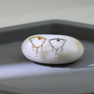 May include: Two earrings, one gold and one silver, each with a curved bar and dangling chains with clear, teardrop-shaped gems. The earrings are displayed on a white, oval stone against a gray background.