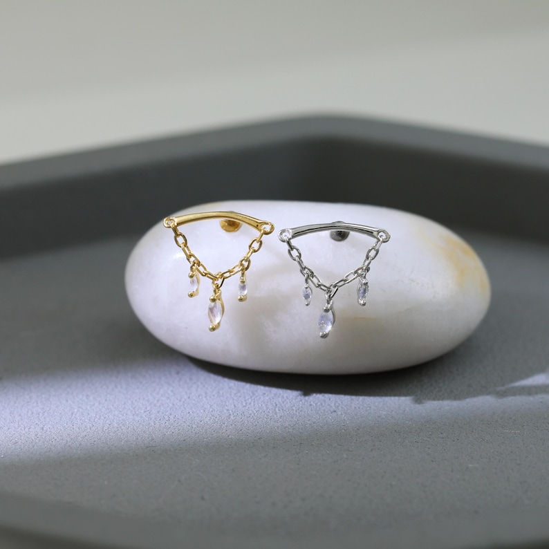 May include: Two earrings, one gold and one silver, each with a curved bar and dangling chains with clear, teardrop-shaped gems. The earrings are displayed on a white, oval stone against a gray background.
