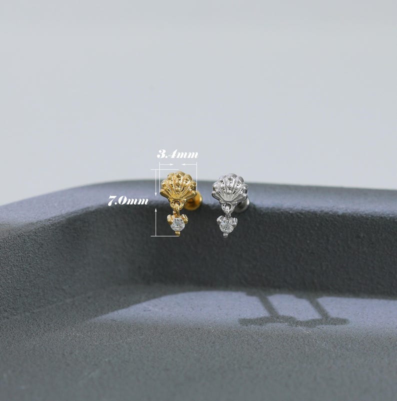 Puede incluir: Dos pendientes de bot&oacute;n, uno dorado y otro plateado, cada uno con una parte superior en forma de concha y una gema transparente colgante. El pendiente dorado mide 7,0 mm de largo y 3,4 mm de ancho.