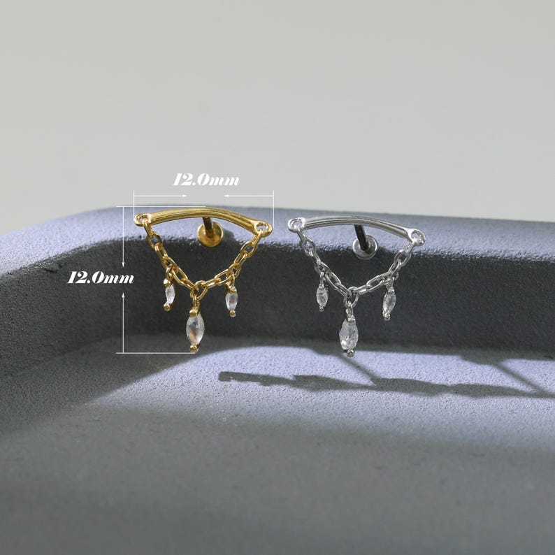 May include: Two earrings, one gold-toned and one silver-toned, each with a curved bar and dangling chains. The chains hold clear, teardrop-shaped gems. Each earring is 12.0mm wide.