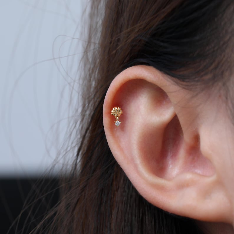 Puede incluir: Un pendiente dorado con un dise&ntilde;o floral y un peque&ntilde;o diamante colgante. El pendiente est&aacute; colocado en la oreja de una persona, mostrando sus delicados detalles y su elegante estilo. El pendiente es un peque&ntilde;o pendiente.