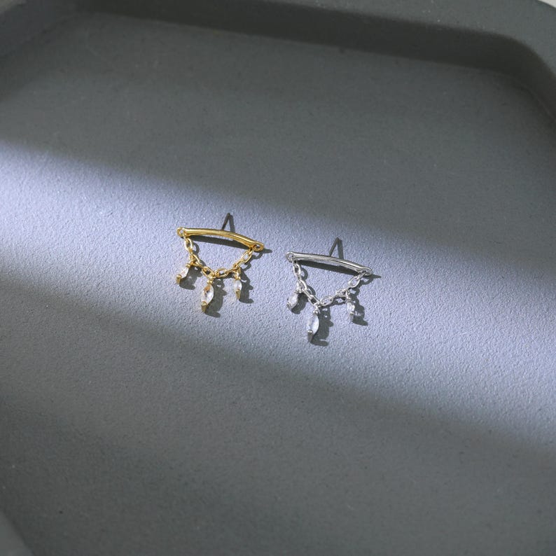 May include: A pair of earrings, one gold-toned and one silver-toned, each featuring a curved bar with dangling clear, teardrop-shaped accents. The earrings are displayed on a gray surface.