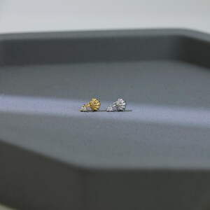 Puede incluir: Dos pendientes de bot&oacute;n, uno dorado y otro plateado, cada uno con un dise&ntilde;o de concha y un peque&ntilde;o acento de gema transparente. Los pendientes se muestran sobre una superficie gris, mostrando sus delicados detalles y colores contrastantes.