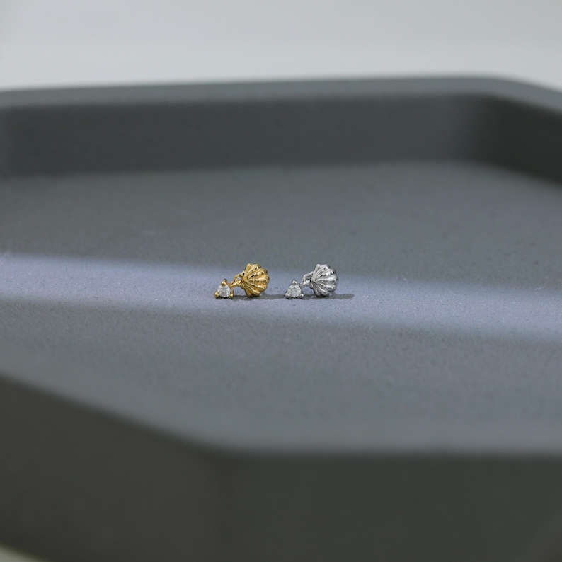 Puede incluir: Dos pendientes de bot&oacute;n, uno dorado y otro plateado, cada uno con un dise&ntilde;o de concha y un peque&ntilde;o acento de gema transparente. Los pendientes se muestran sobre una superficie gris, mostrando sus delicados detalles y colores contrastantes.