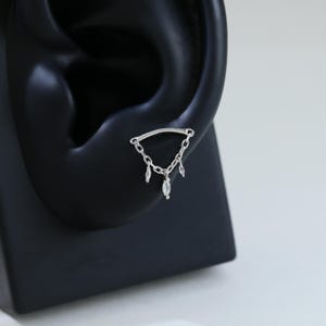 May include: A silver ear piercing featuring a curved bar connected to a chain with three dangling, teardrop-shaped clear gemstones. The jewelry is displayed on a black ear model against a white background.