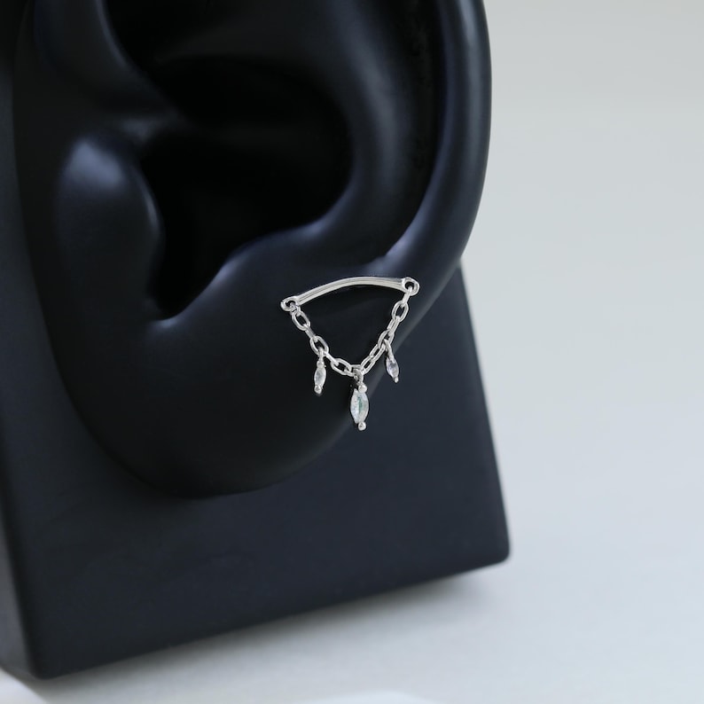 May include: A silver ear piercing featuring a curved bar connected to a chain with three dangling, teardrop-shaped clear gemstones. The jewelry is displayed on a black ear model against a white background.