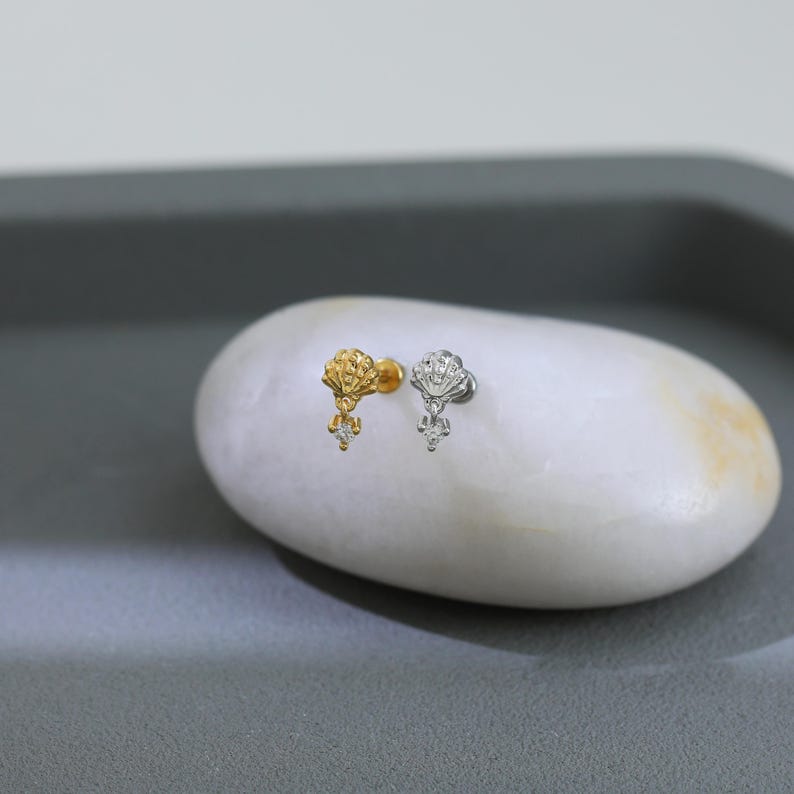 Puede incluir: Dos pendientes de bot&oacute;n, uno dorado y otro plateado, cada uno con un dise&ntilde;o de concha y una gema transparente colgante. Los pendientes se muestran sobre una piedra ovalada blanca sobre un fondo gris.