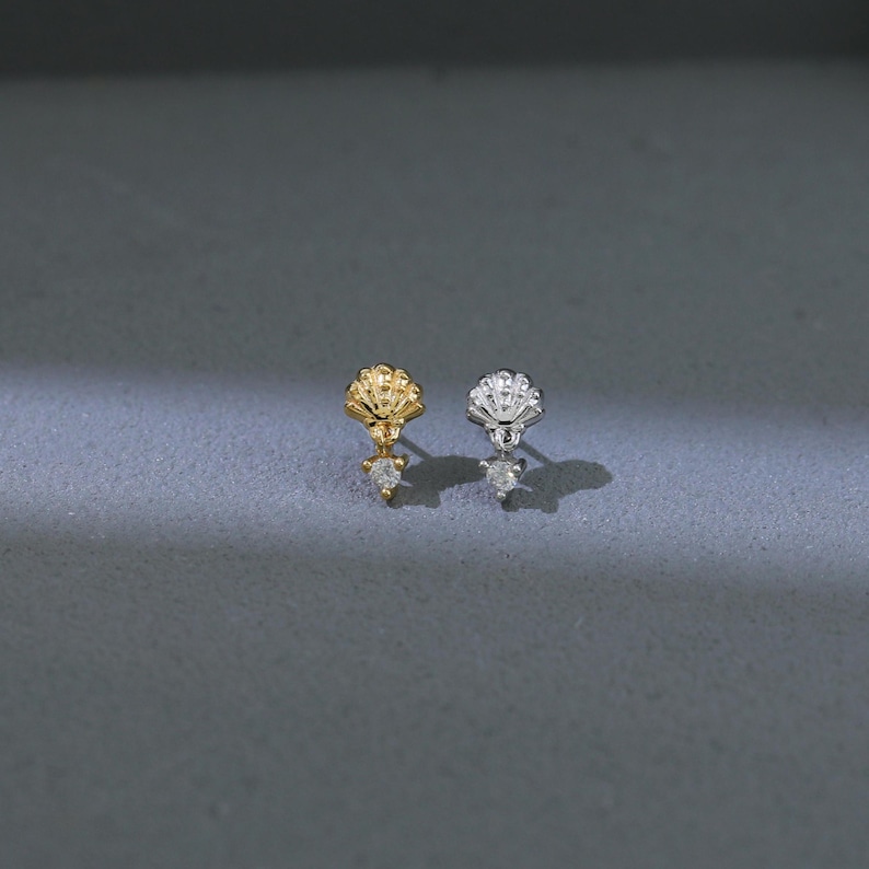 Puede incluir: Dos pendientes de bot&oacute;n, uno dorado y otro plateado, cada uno con un dise&ntilde;o de concha y una piedra transparente en forma de l&aacute;grima colgante. Los pendientes se muestran sobre una superficie gris.