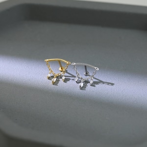 May include: Two pieces of jewelry, one gold and one silver, each featuring a curved bar with dangling teardrop-shaped charms. The jewelry is designed for body piercings, with a small post on the back for insertion.