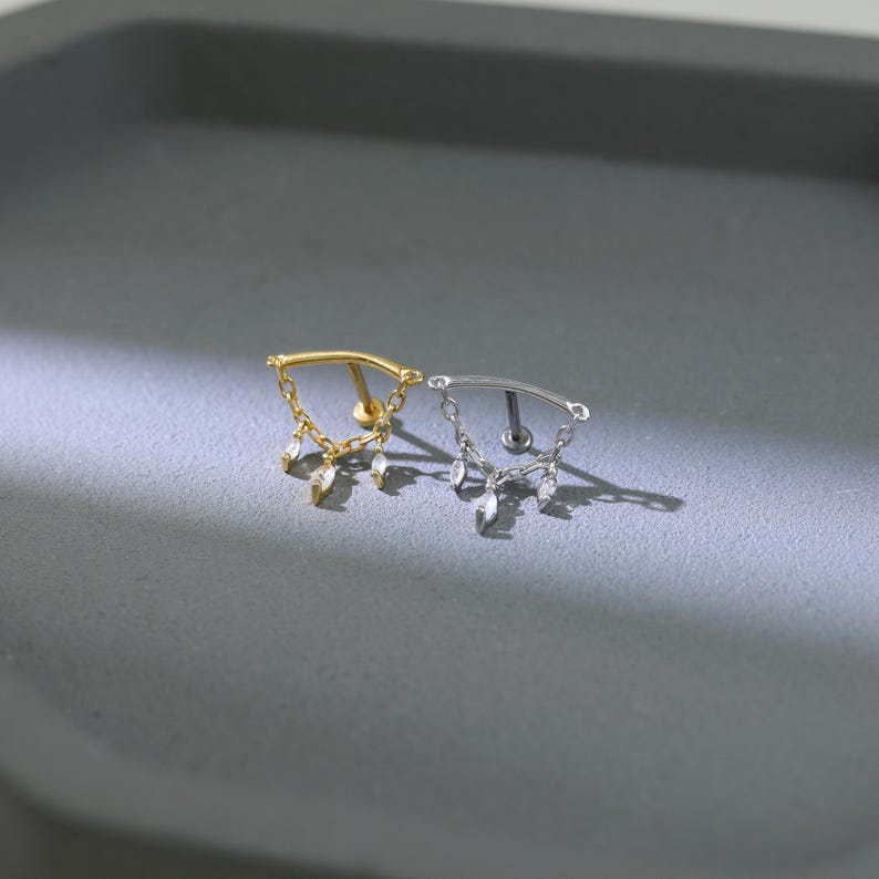 May include: Two pieces of jewelry, one gold and one silver, each featuring a curved bar with dangling teardrop-shaped charms. The jewelry is designed for body piercings, with a small post on the back for insertion.