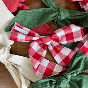 Op de afbeelding: Een verzameling stoffen haarstrikken met diverse ontwerpen, waaronder een rood-wit vichy-ruitpatroon, effen bosgroen en effen crème. Elke strik is geconstrueerd met lange, puntige stoffen uiteinden en een centraal knoopdetail.