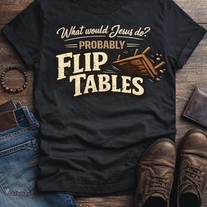 May include: Black t-shirt with the text "What would Jesus do? Probably Flip Tables" in a distressed font. The shirt is laid out with jeans, a bracelet, a wallet, and brown leather shoes on a wooden surface.