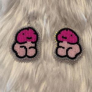 Könnte beinhalten: Ein Paar Perlenohrringe mit einem verspielten Design. Jeder Ohrring zeigt eine rosa und schwarze Perlenform mit einem Perlenrand. Die Ohrringe hängen an silberfarbenen Haken und sind vor einem flauschigen, weißen Hintergrund platziert.