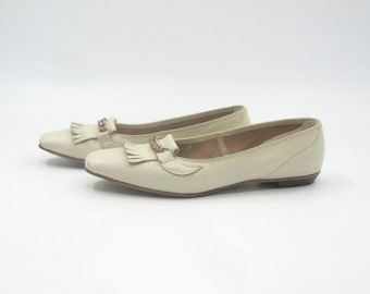 Puede incluir: Un par de mocasines de cuero color crema con detalles de borlas y hebillas. Los zapatos tienen un tacón bajo y un diseño clásico. Los mocasines se muestran sobre un fondo blanco. Adecuados para el uso diario.